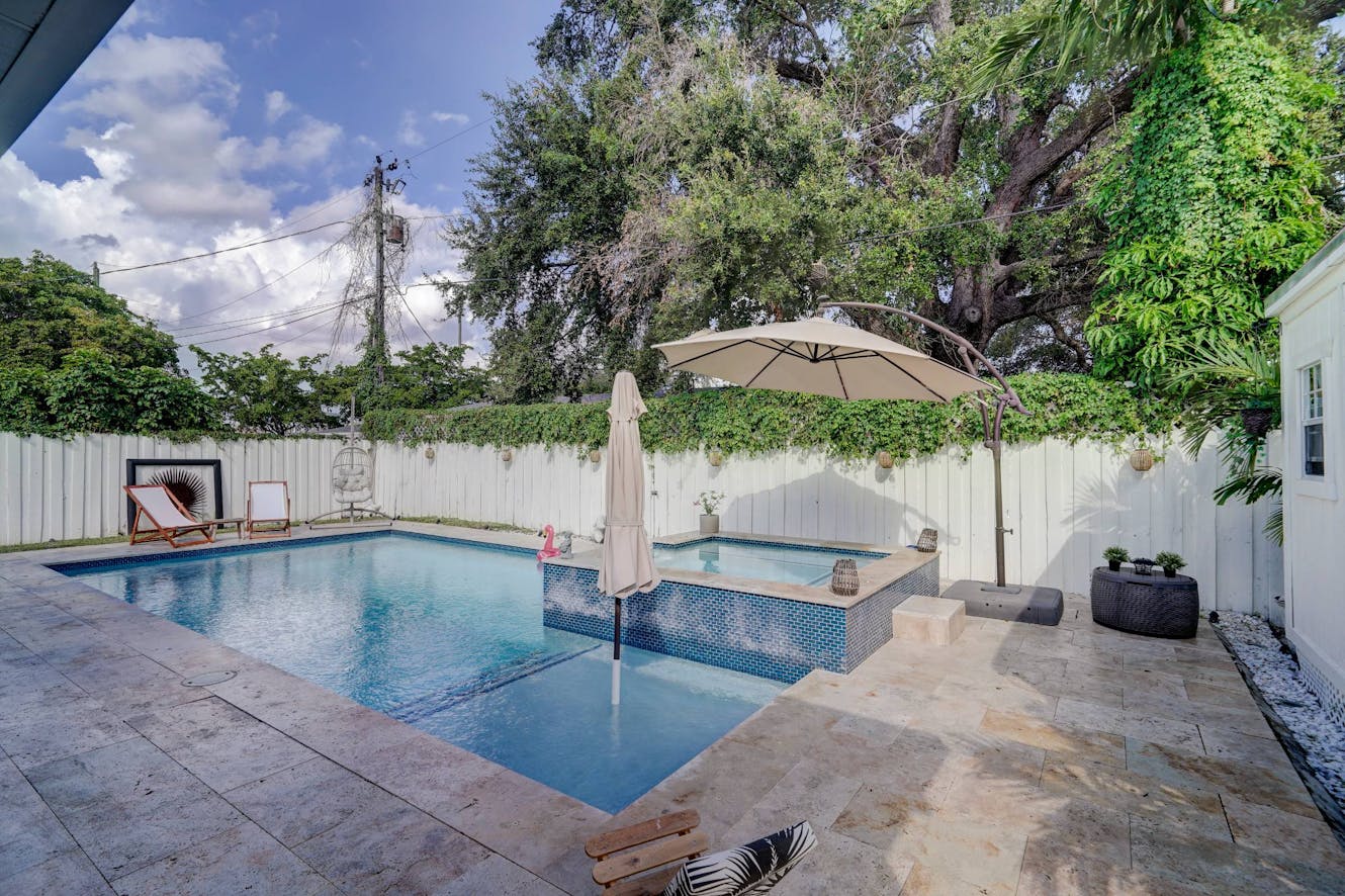 Backyard pool with blue tile, raised hot tub, lounge chairs, patio umbrella, white privacy fence, and mature trees