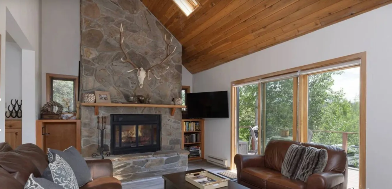 Living room with vaulted wood ceiling, stone fireplace with antler mount, leather sofa, large windows with tree views, and wall-mounted TV