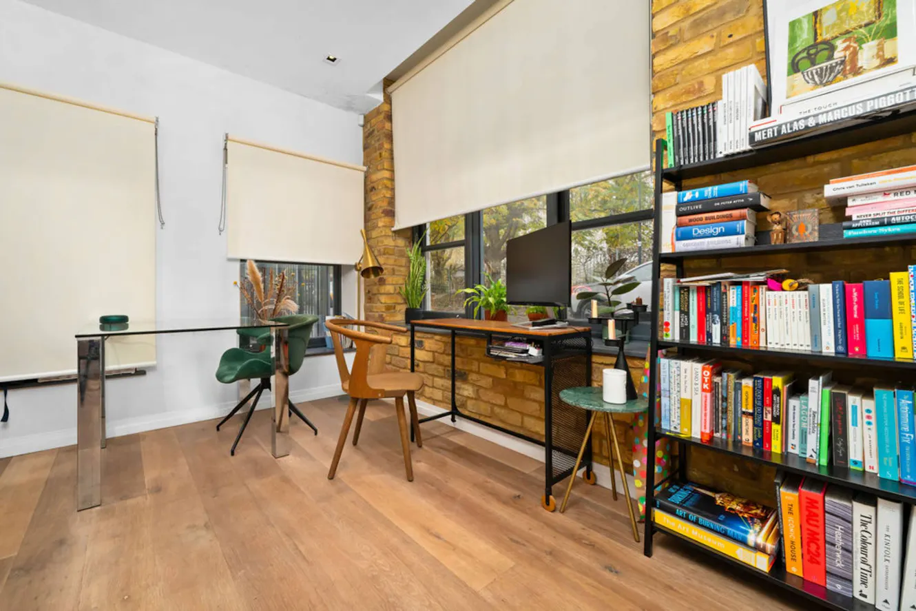 Workspace with glass desk, wooden chairs, exposed brick wall, large windows with natural light, and tall bookshelf filled with colorful books