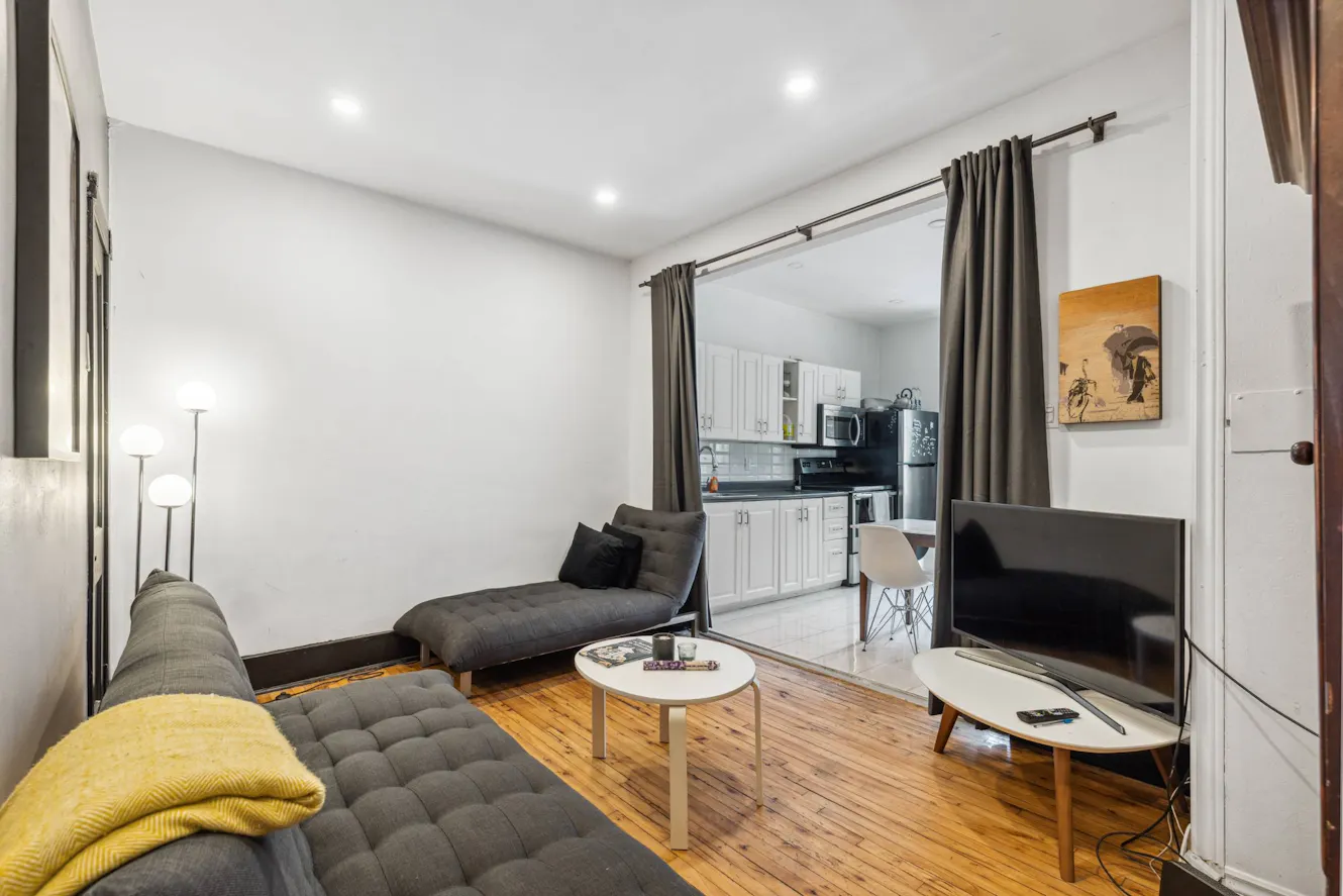 Living room with gray tufted sofa, chaise lounge, wooden coffee tables, floor lamp, flat-screen TV, and view into white kitchen