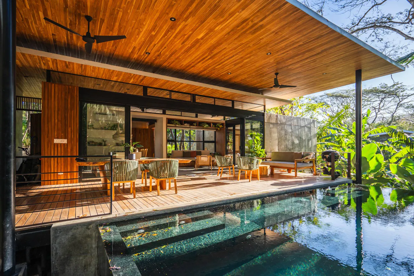 Covered patio with wooden deck, outdoor dining furniture, ceiling fans, and adjacent swimming pool with tropical landscaping