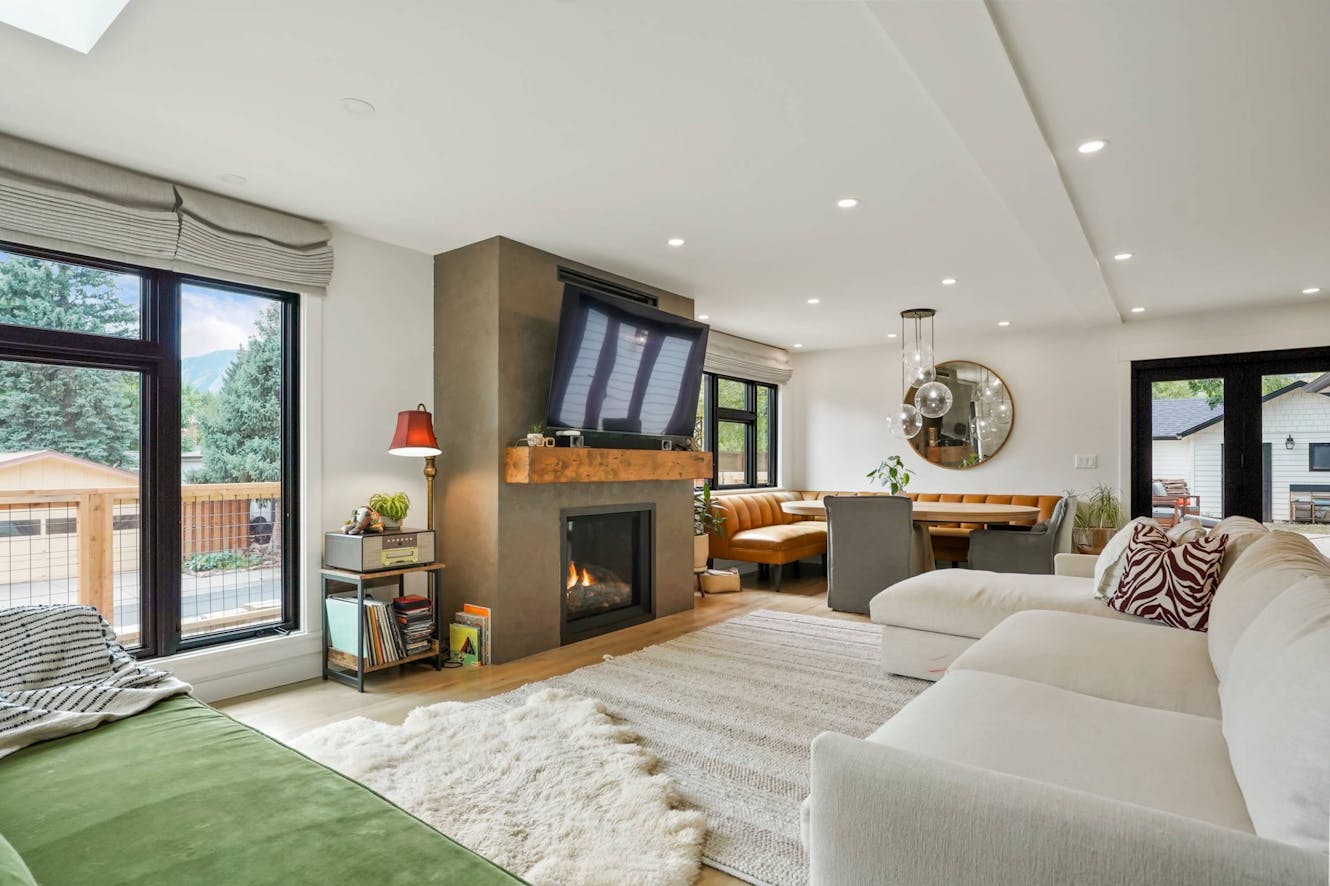 Living room with white sectional sofa, modern fireplace with wood mantel, wall-mounted TV, large windows with mountain views, and open dining area with orange leather seating