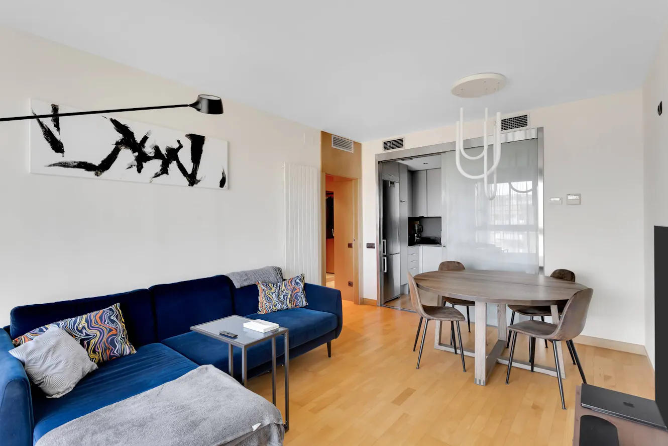 Living room and dining area with blue sectional sofa, light wood flooring, round dining table with four chairs, modern white pendant light, and view into kitchen