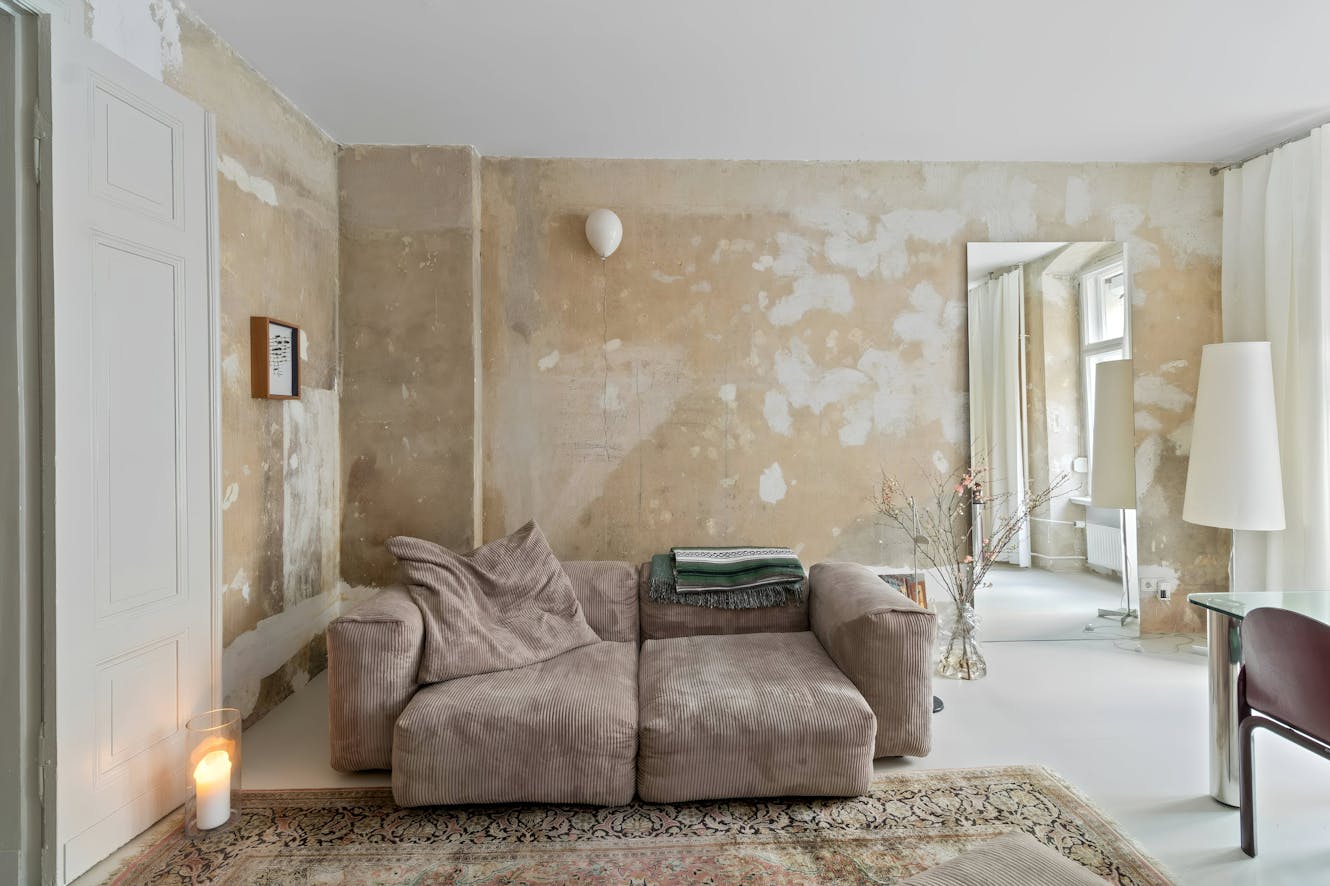 Living room with tan modular sofa, distressed plaster walls, large mirror, patterned area rug, and white floor lamp