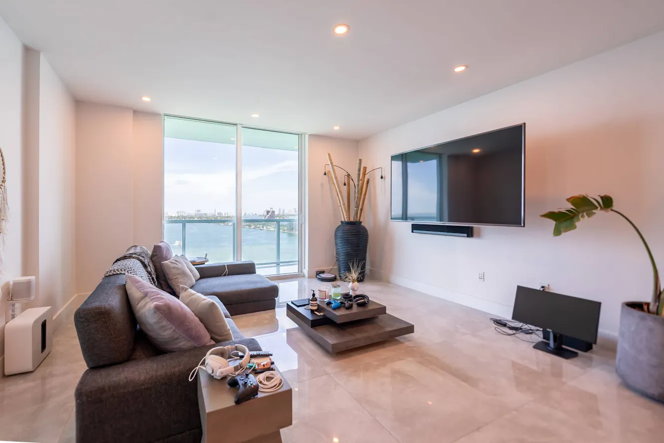 Living room with gray sectional sofa, floor-to-ceiling windows with ocean view, wall-mounted TV, modern coffee table, and recessed lighting