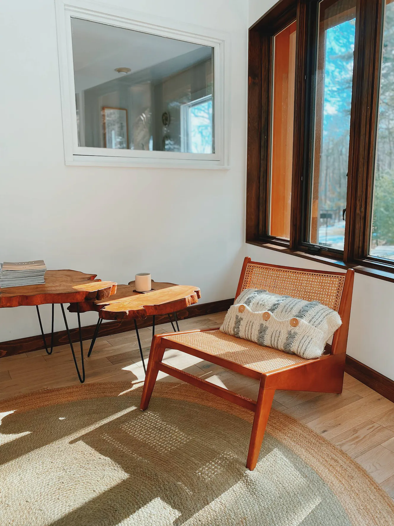 Living room with cane accent chair, live edge wood coffee table, white walls, large dark wood-framed window with natural light, and patterned area rug