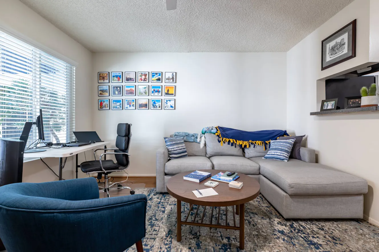 Living room with gray sectional sofa, blue accent chair, round coffee table, workspace with desk and office chair, large window with blinds, and gallery wall of framed photos