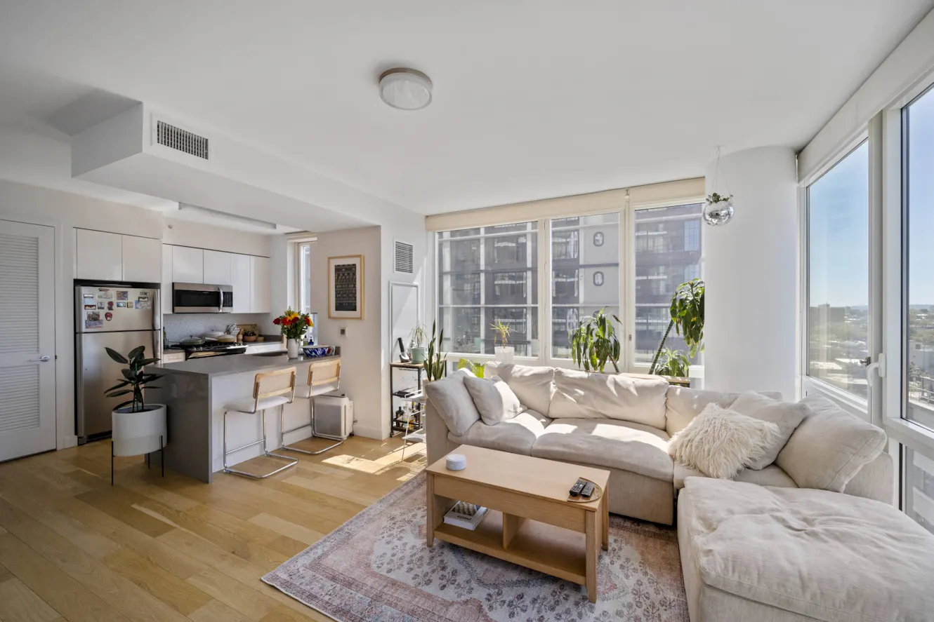 Open-concept living room and kitchen with beige sectional sofa, wooden coffee table, floor-to-ceiling windows with city views, white kitchen with breakfast bar and stools