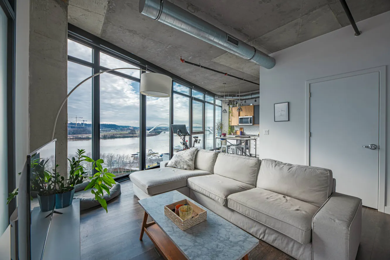 Living room with gray sectional sofa, floor-to-ceiling windows with river view, exposed concrete ceiling, arc floor lamp, and open kitchen area