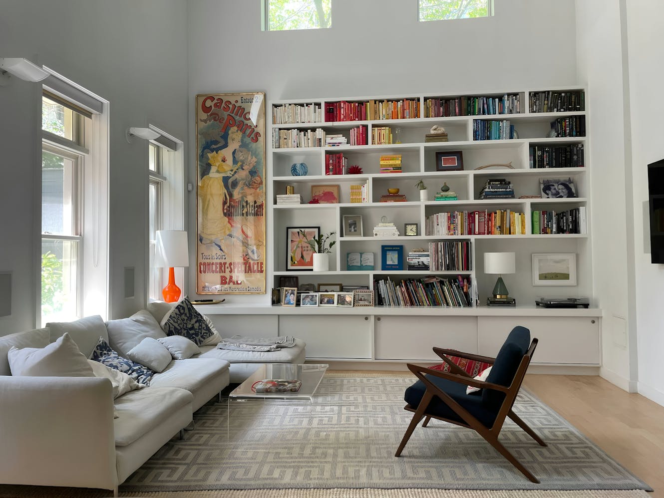 Living room with white sectional sofa, built-in bookshelf with colorful books, glass coffee table, mid-century modern chair, and large windows with natural light