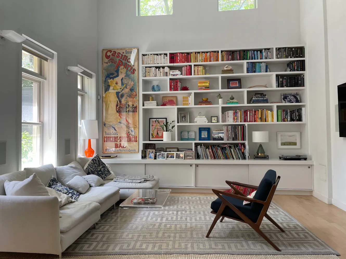 Living room with white sectional sofa, built-in bookshelf with colorful books, glass coffee table, mid-century modern chair, and large windows with natural light