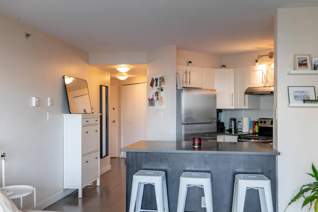 Kitchen with white cabinets, stainless steel appliances, gray island with three white bar stools, and open layout with entry hallway visible