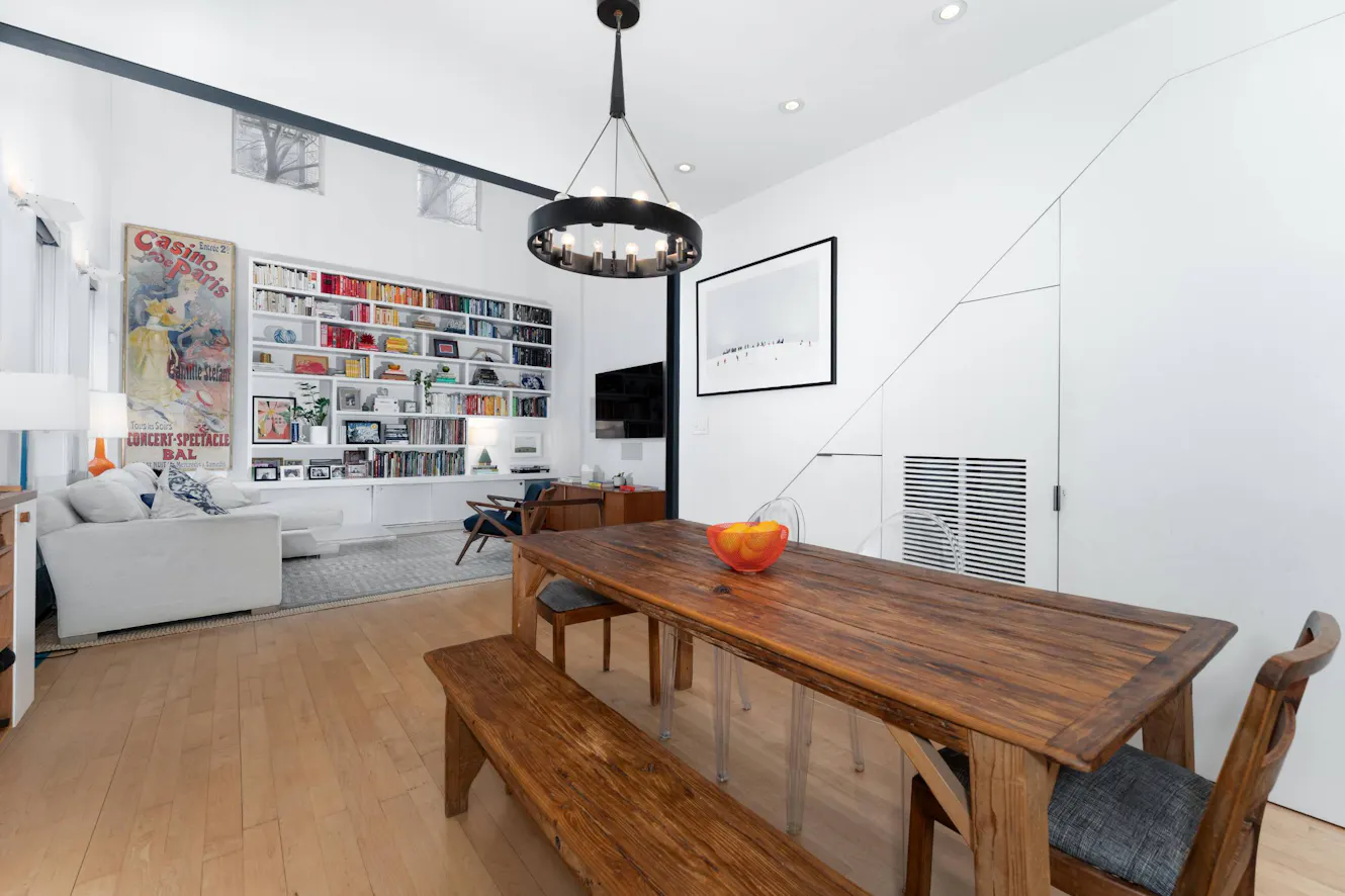 Open-concept living and dining area with wooden dining table and benches, white sofa, built-in bookshelf, modern chandelier, and light wood flooring