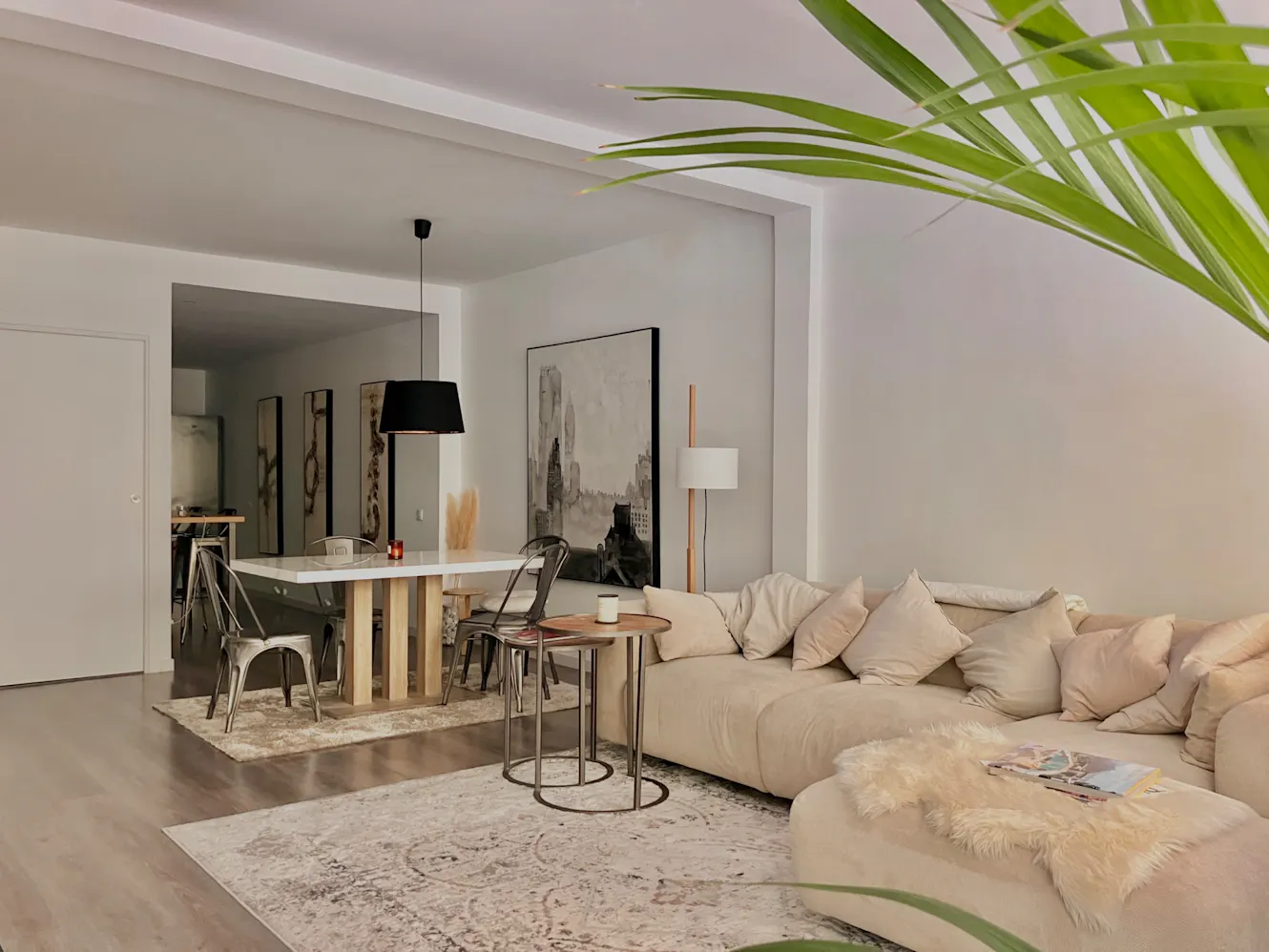 Living room with beige sectional sofa, nesting coffee tables, dining table with metal chairs, black pendant light, and abstract artwork on white walls
