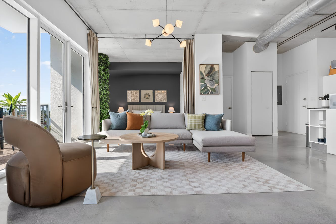 Modern living room with gray sectional sofa, round wooden coffee table, exposed concrete ceiling, floor-to-ceiling windows, and view of bedroom with dark accent wall