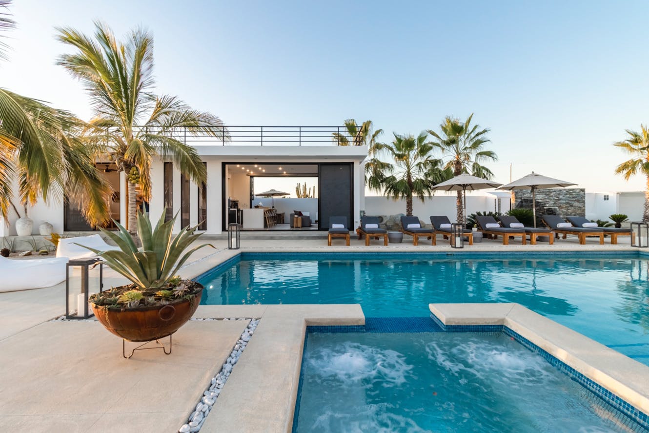 Pool area with hot tub, lounge chairs, palm trees, modern white home with open glass doors, and roof deck with railing
