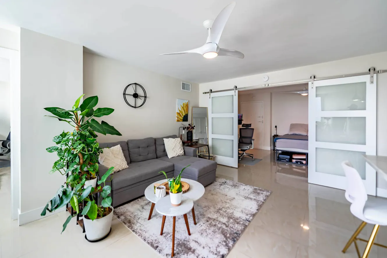 Living room with gray sectional sofa, white round coffee table, large potted plant, ceiling fan, and frosted glass sliding barn doors leading to bedroom