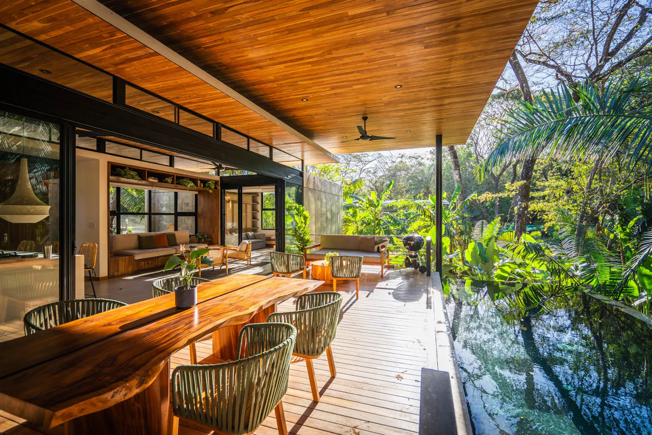 Covered patio with wooden deck, dining table with chairs, ceiling fan, pool along edge, and tropical garden views with open-air living space beyond