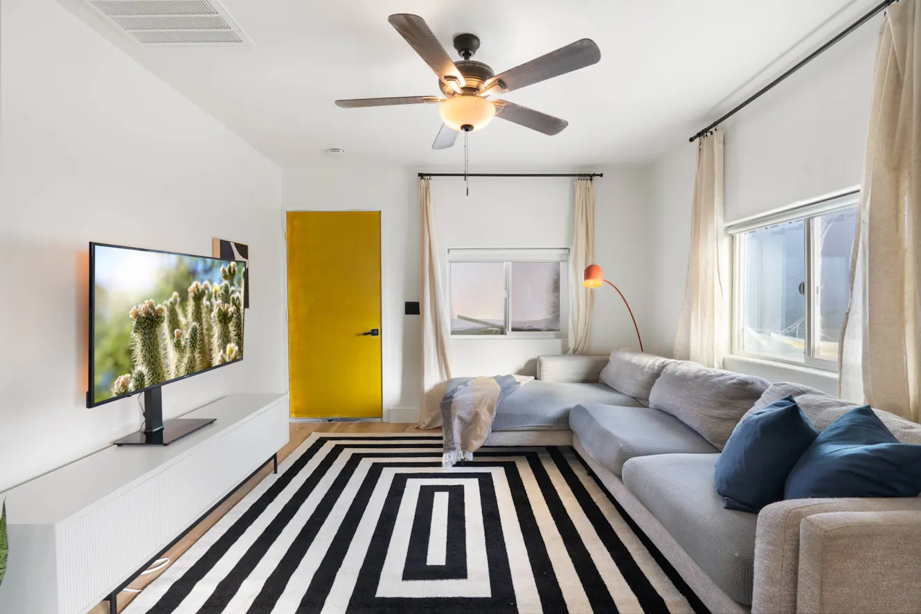 Living room with gray sectional sofa, wall-mounted TV, ceiling fan, geometric black and white rug, and windows with natural light