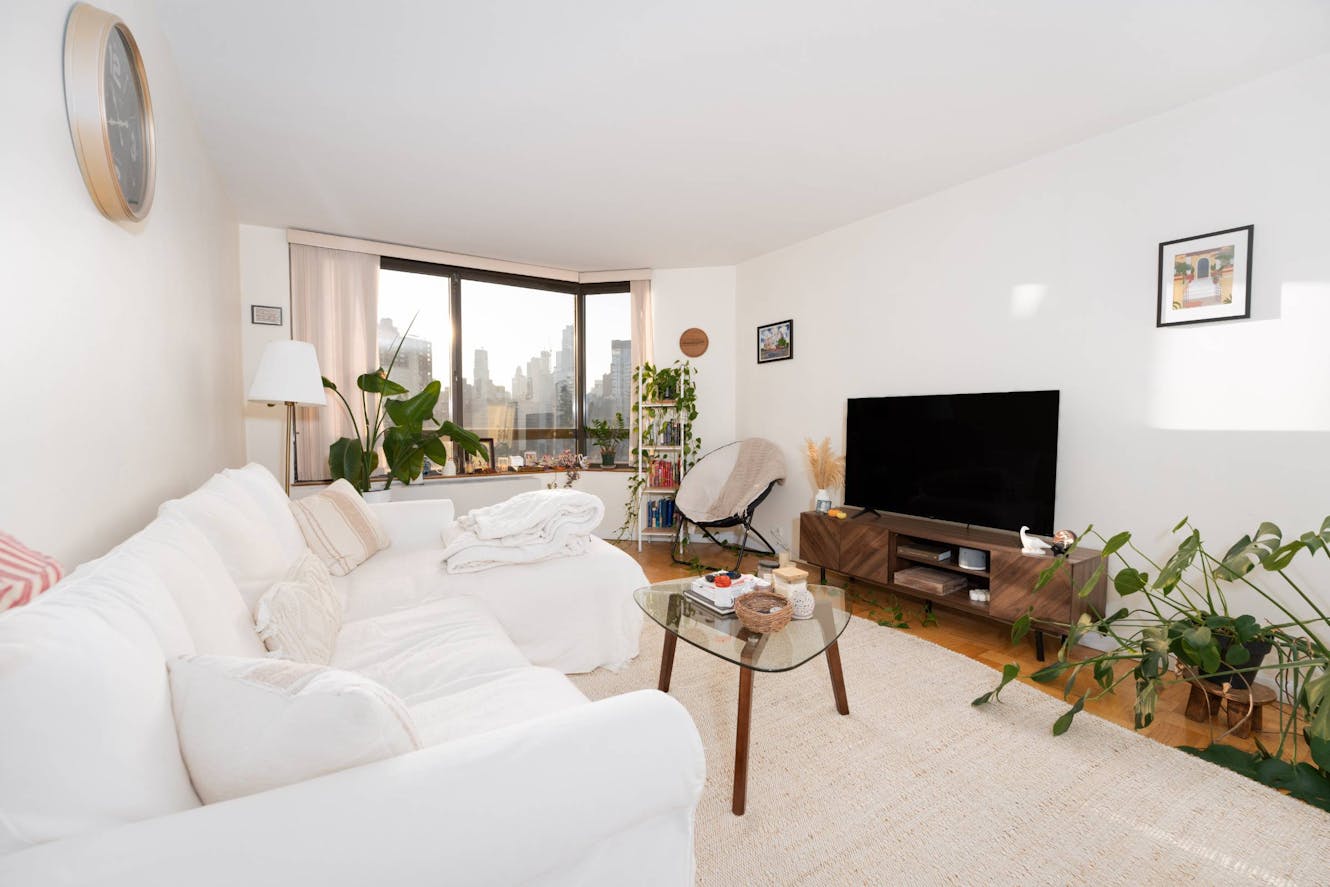 Living room with white sectional sofa, glass coffee table, wooden TV stand with flat-screen TV, large corner windows with city skyline view, and indoor plants