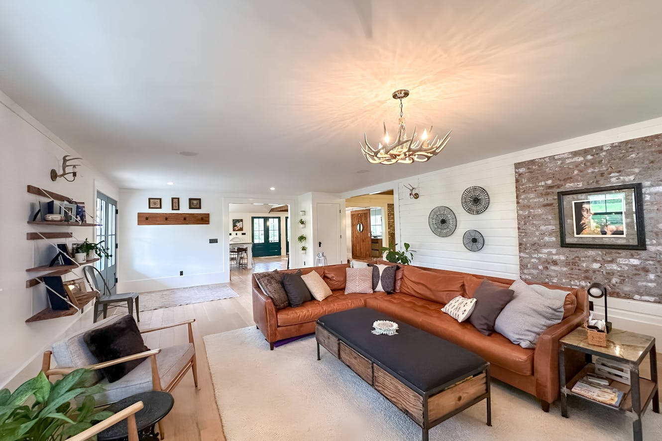 Living room with brown leather sectional sofa, antler chandelier, exposed brick accent wall, white shiplap walls, and open floor plan leading to other rooms