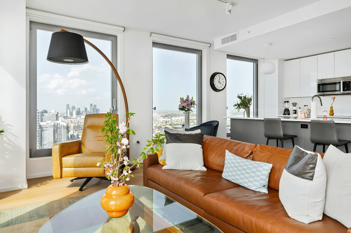 Living room with tan leather sofa, glass coffee table, arc floor lamp, large windows with city skyline views, and open kitchen with white cabinets and bar seating