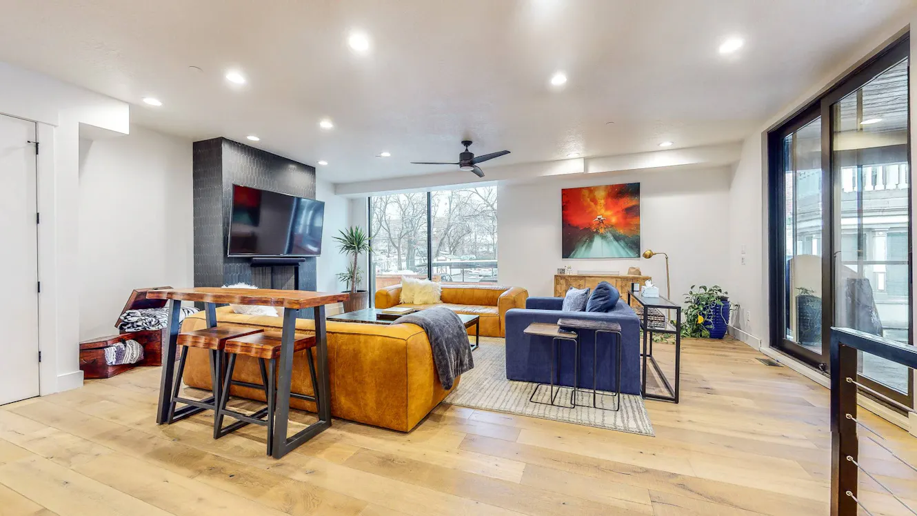 Living room with tan leather sectional sofa, blue accent chairs, wall-mounted TV above fireplace, large windows with natural light, and light wood flooring