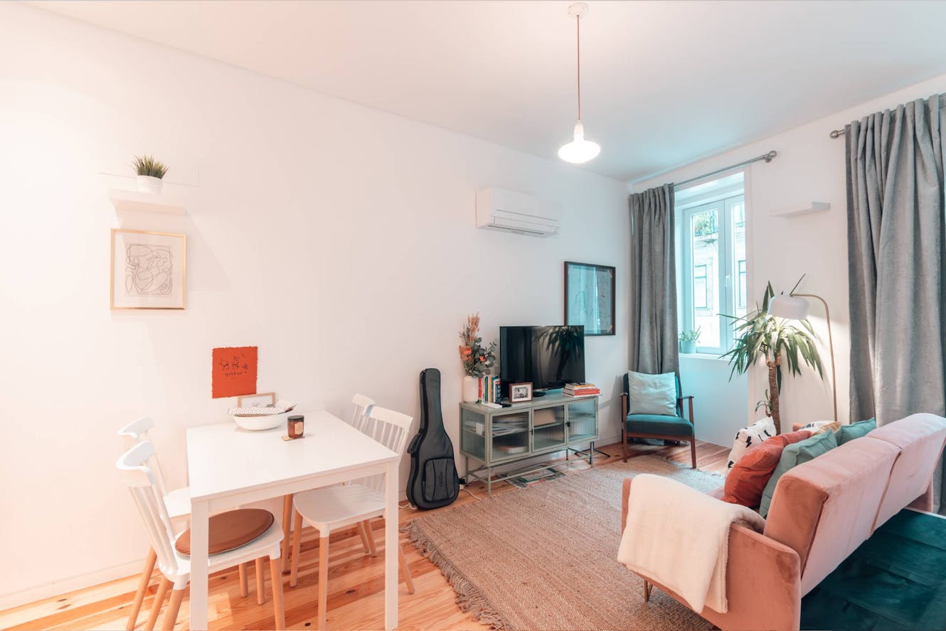 Living room with pink sofa, white dining table with chairs, TV on media console, gray curtains, window with natural light, and wall-mounted AC unit