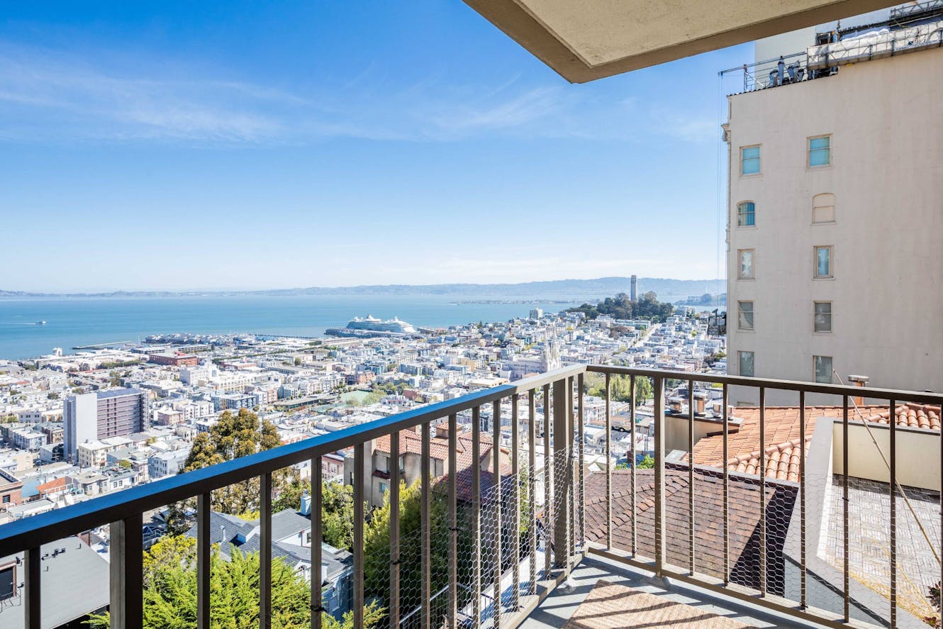 Patio balcony with metal railing overlooking city skyline, bay, and distant mountains under clear blue sky