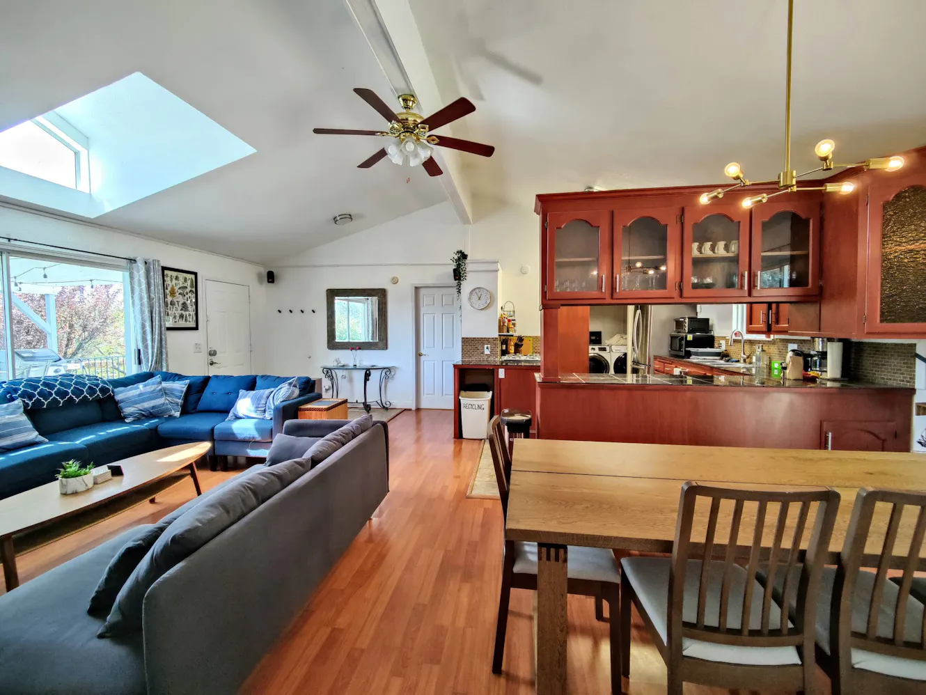 Open concept living room and kitchen with vaulted ceiling, skylight, blue sectional sofa, dining table, and cherry wood cabinets