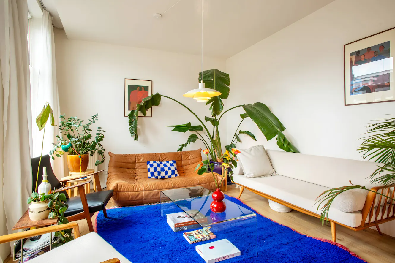 Living room with tan leather sofa, white loveseat, glass coffee table on bright blue rug, large windows with natural light, and tropical plants