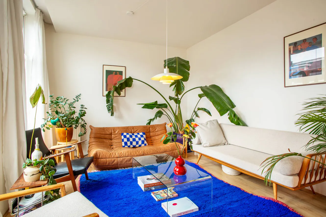 Living room with tan leather sofa, white loveseat, glass coffee table on bright blue rug, large windows with natural light, and tropical plants