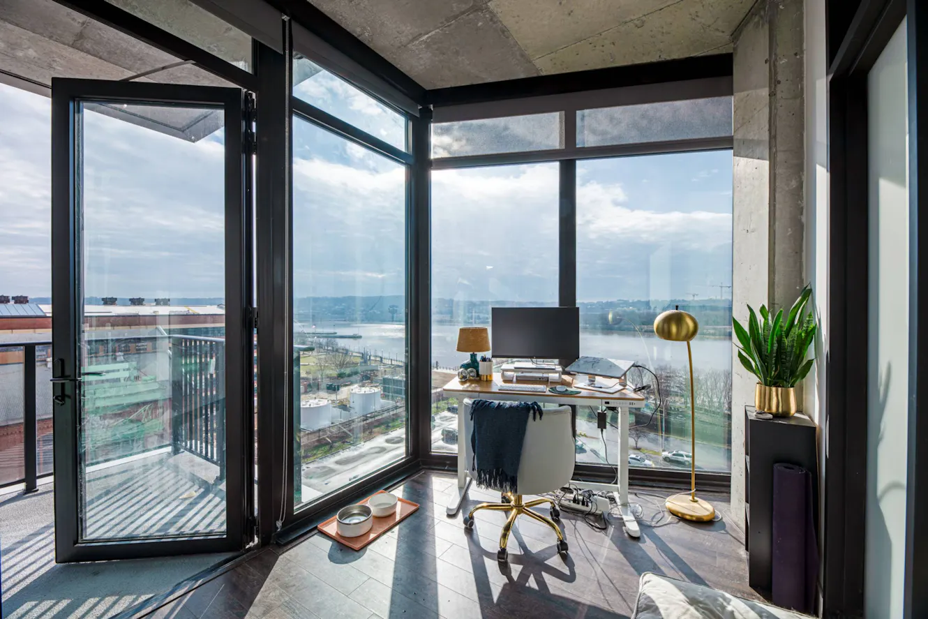 Workspace with glass desk, white office chair, dual monitors, floor-to-ceiling windows with water view, exposed concrete ceiling, and balcony access