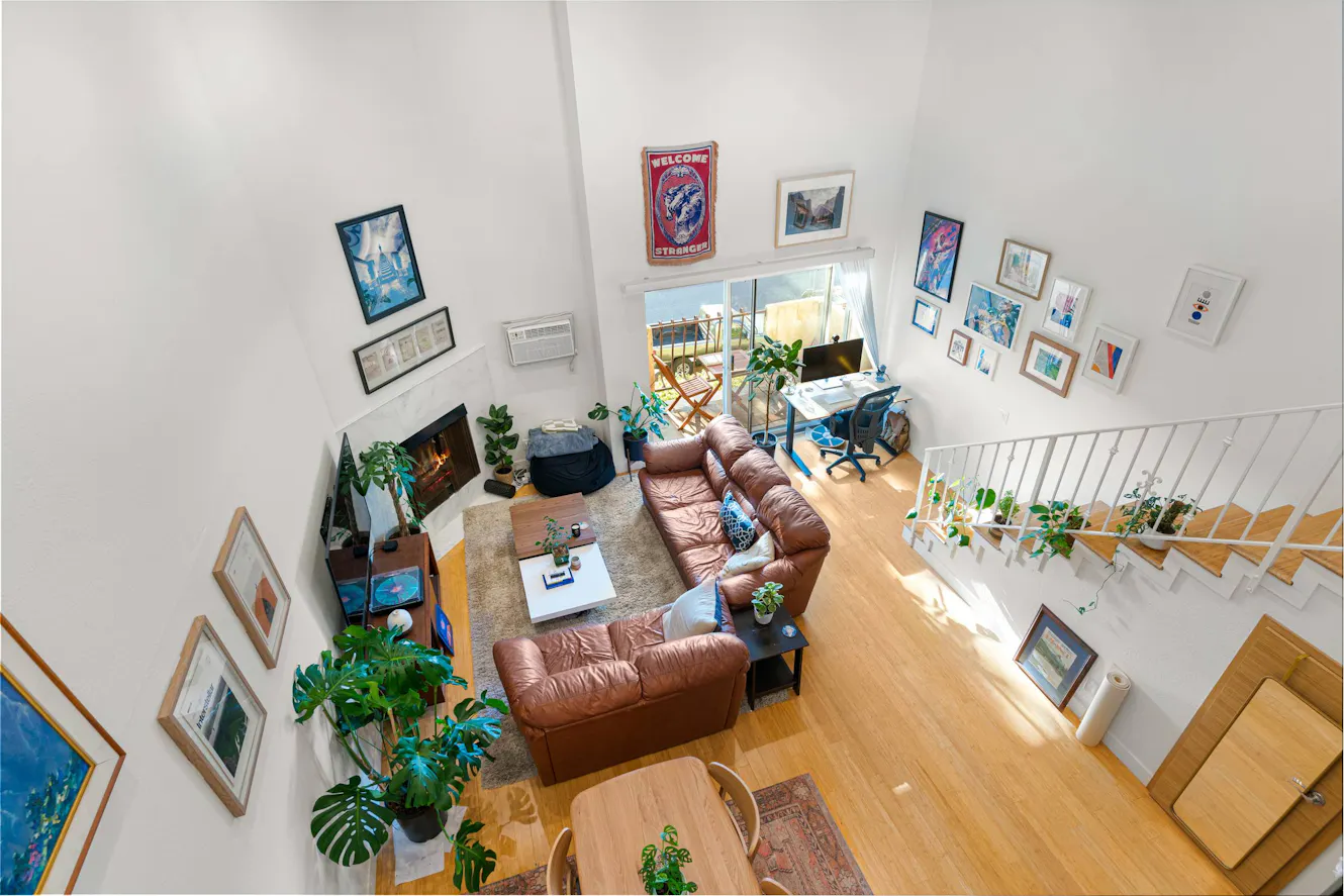 Living room with high ceilings, brown leather sectional sofa, workspace with desk and office chair, large windows with natural light, and gallery wall with framed artwork