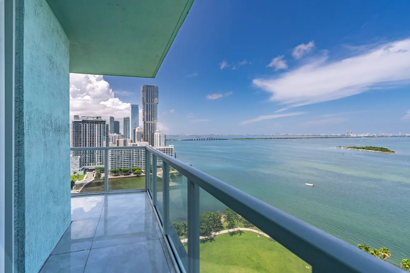 Balcony with glass railing overlooking waterfront bay, city skyline with high-rise buildings, and green park area below