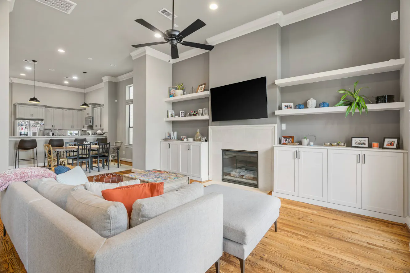Open concept living room with gray sectional sofa, wall-mounted TV, built-in fireplace with white cabinetry, floating shelves, ceiling fan, and view into kitchen and dining area