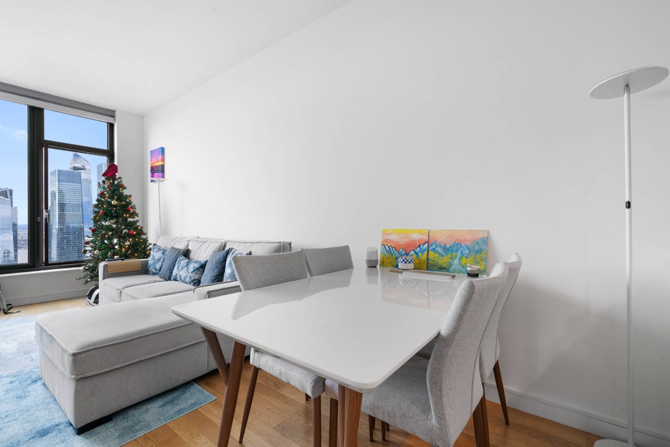 Living room with gray sectional sofa, white dining table with chairs, large windows with city views, and decorated Christmas tree