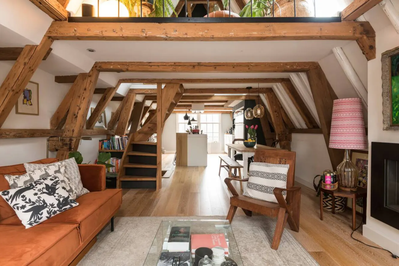 Living room with exposed wooden beams, orange sofa, glass coffee table, wooden chair, loft area above, and view into kitchen
