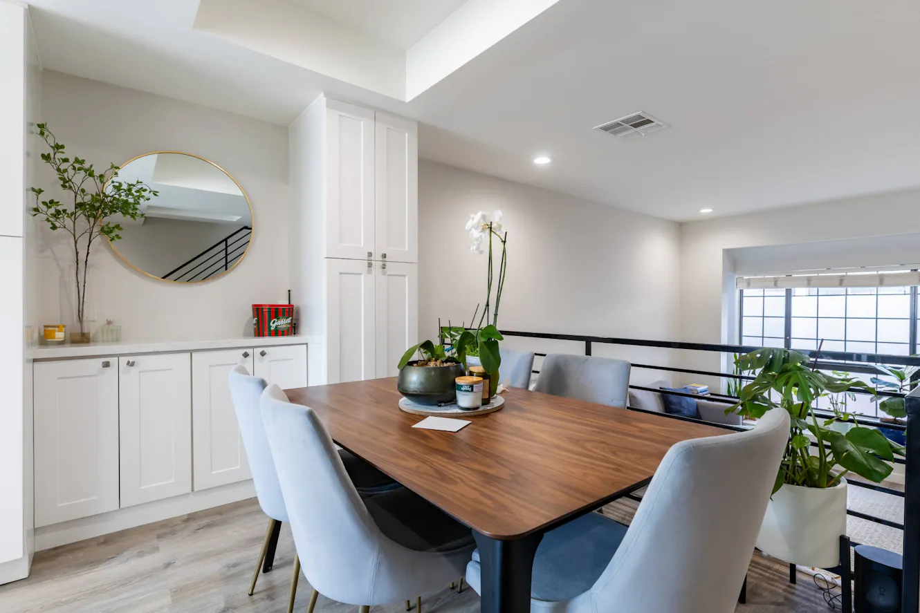 Dining room with wooden table, upholstered chairs, white built-in cabinets with round mirror, and black metal railing overlooking lower level