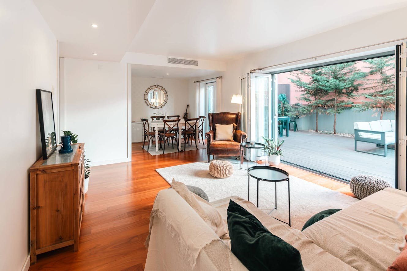 Living room with beige sofa, wooden flooring, leather armchair, floor lamp, and large sliding glass doors opening to outdoor patio with trees