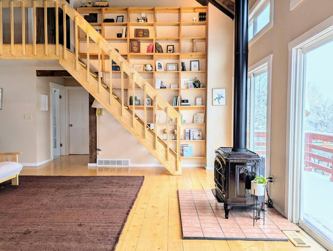 Living room with wooden staircase to loft, floor-to-ceiling built-in bookshelf, wood-burning stove, light wood flooring, and large windows with natural light