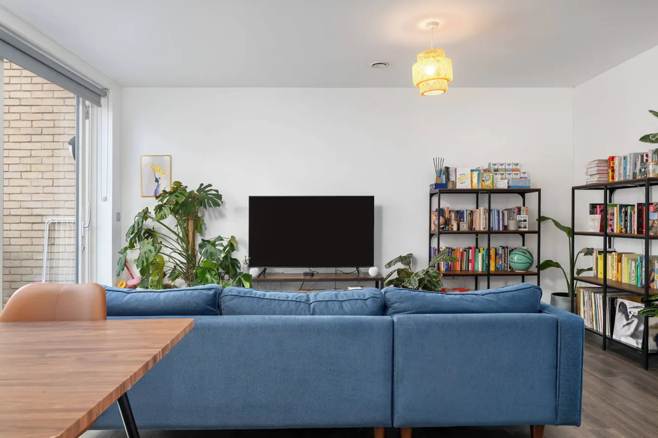 Living room with blue sectional sofa, wall-mounted TV, bookshelves, indoor plants, sliding glass door, and woven pendant light