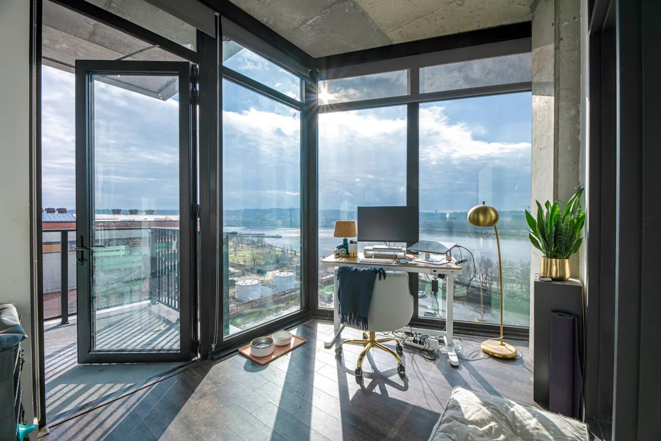 Modern workspace with glass desk, white office chair, floor-to-ceiling windows with waterfront view, exposed concrete ceiling, and access to balcony