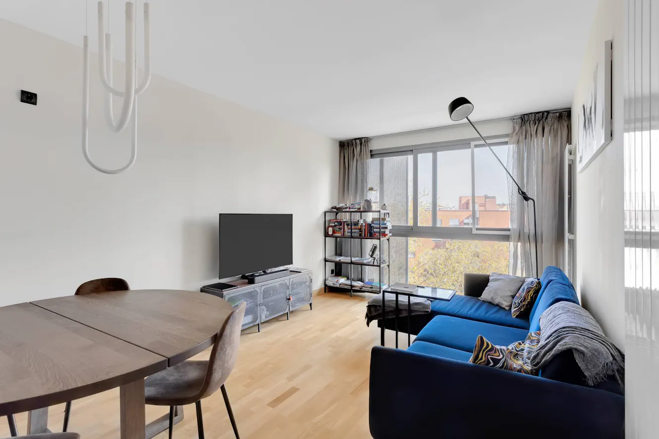 Living room with blue sofa, dining table, wall-mounted TV on media console, floor lamp, shelving unit, and large windows with city view