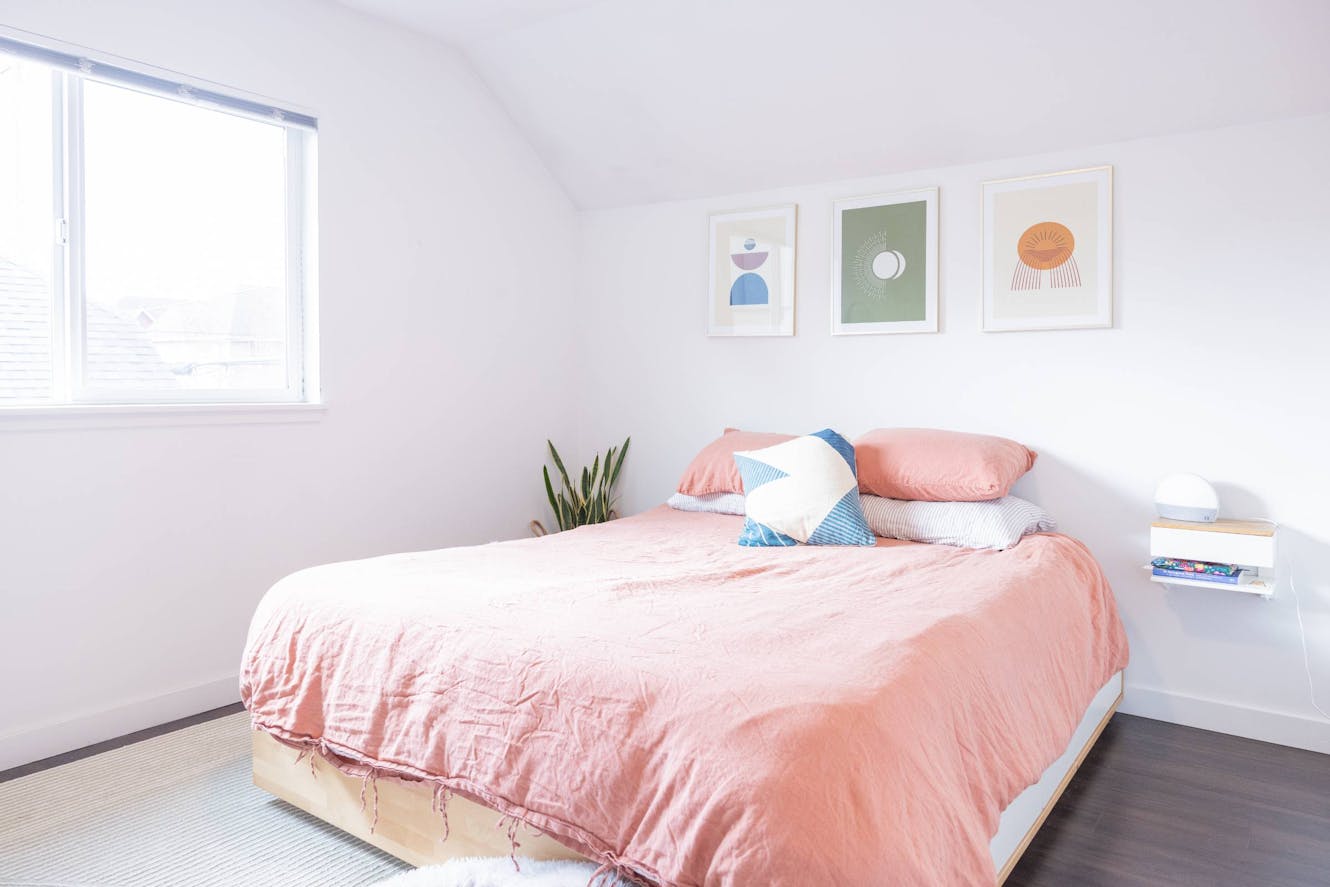 Bedroom with platform bed, pink bedding, white walls, window with natural light, wall art, and floating nightstand