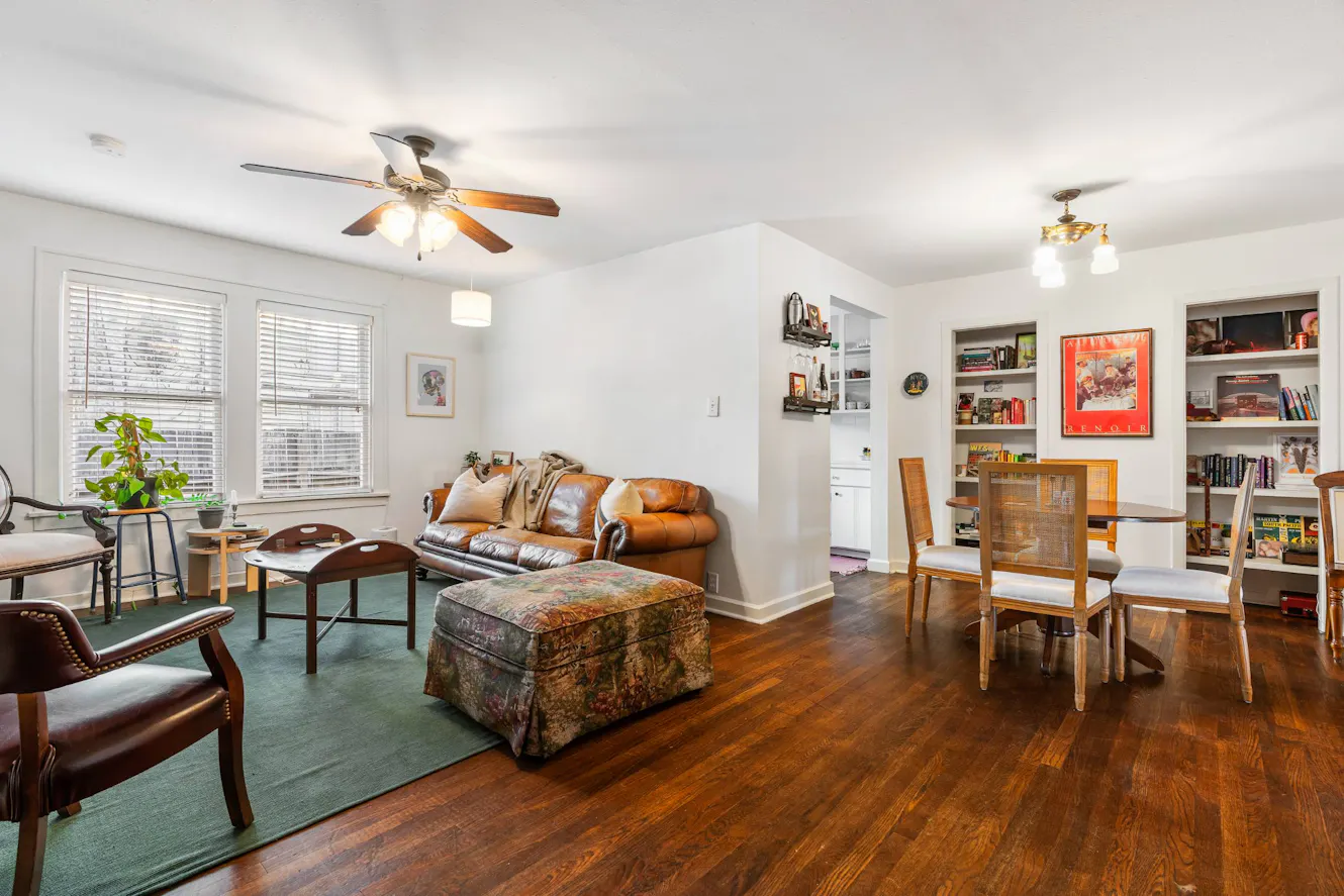 Living room with brown leather sofa, dining table with chairs, built-in white bookshelves, hardwood floors, ceiling fan, and two large windows with blinds