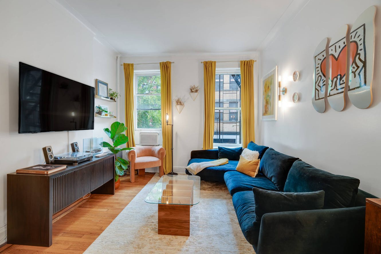 Living room with blue velvet sectional sofa, glass coffee table, wall-mounted TV, two windows with yellow curtains, and hardwood floors