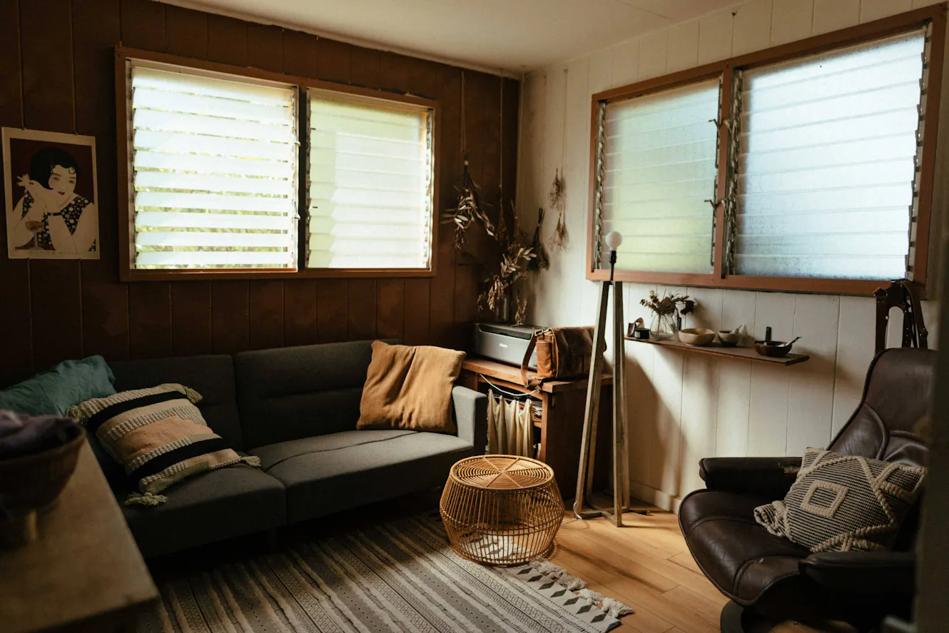 Living room with dark green sofa, leather chair, wicker ottoman, floor lamp, wood-paneled walls, and louvered windows with natural light