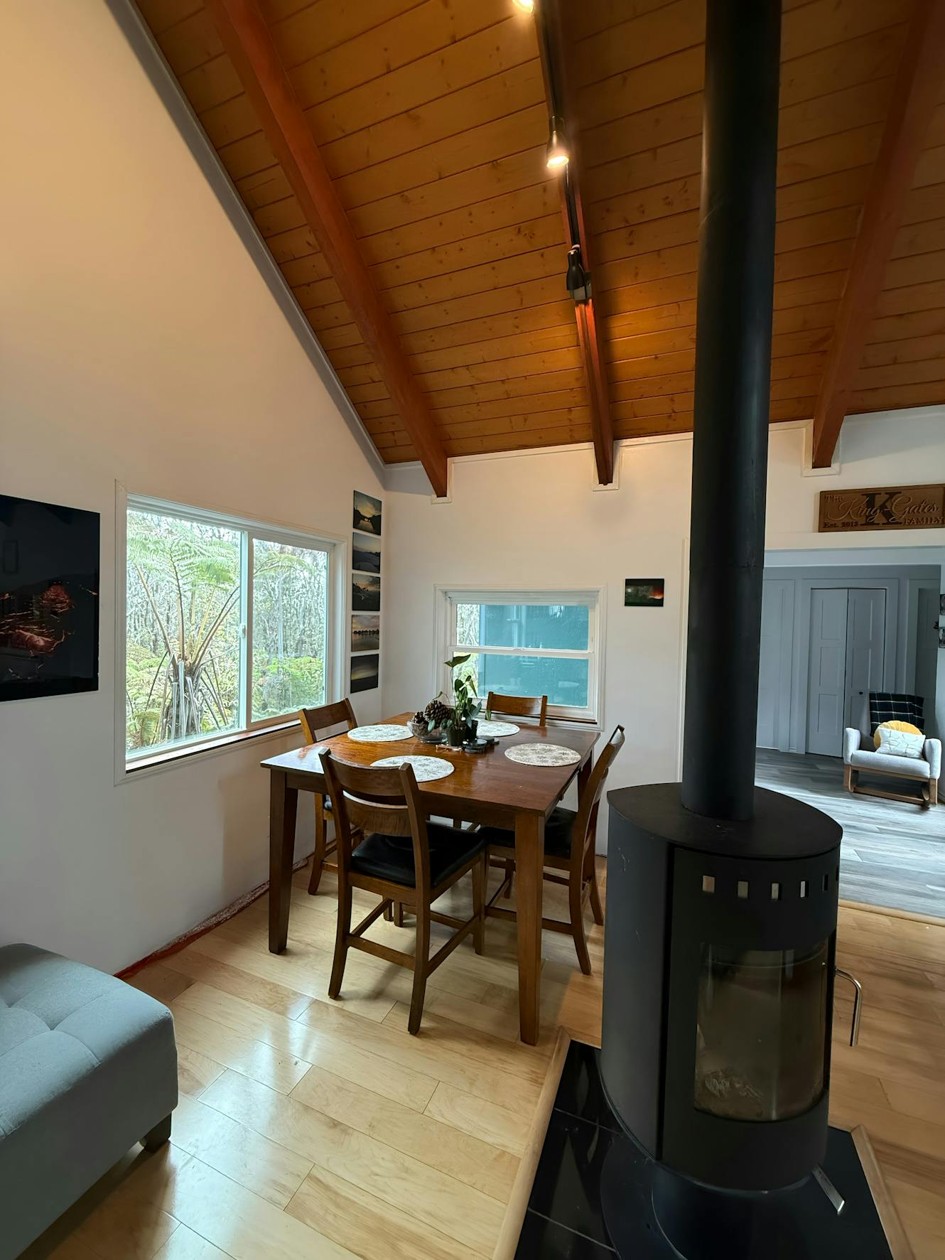 Dining room with wooden table and chairs, vaulted wood ceiling, large windows with forest view, and black wood-burning stove