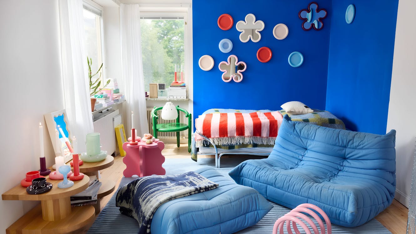 Living room with bright blue accent wall, blue floor cushions, striped daybed, large windows with natural light, and colorful wall decorations